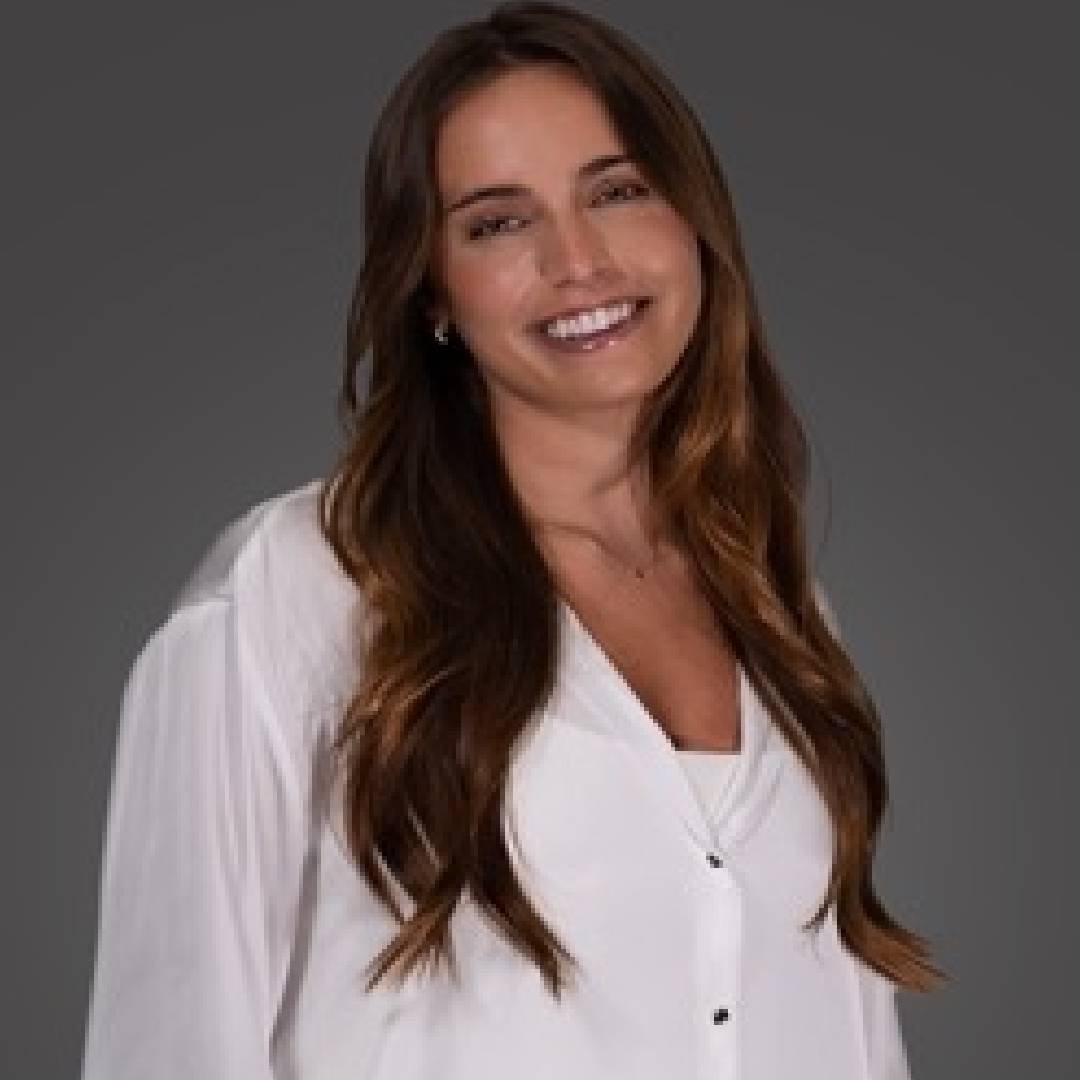Smiling woman with long brown hair wearing a white blouse against a gray background.
