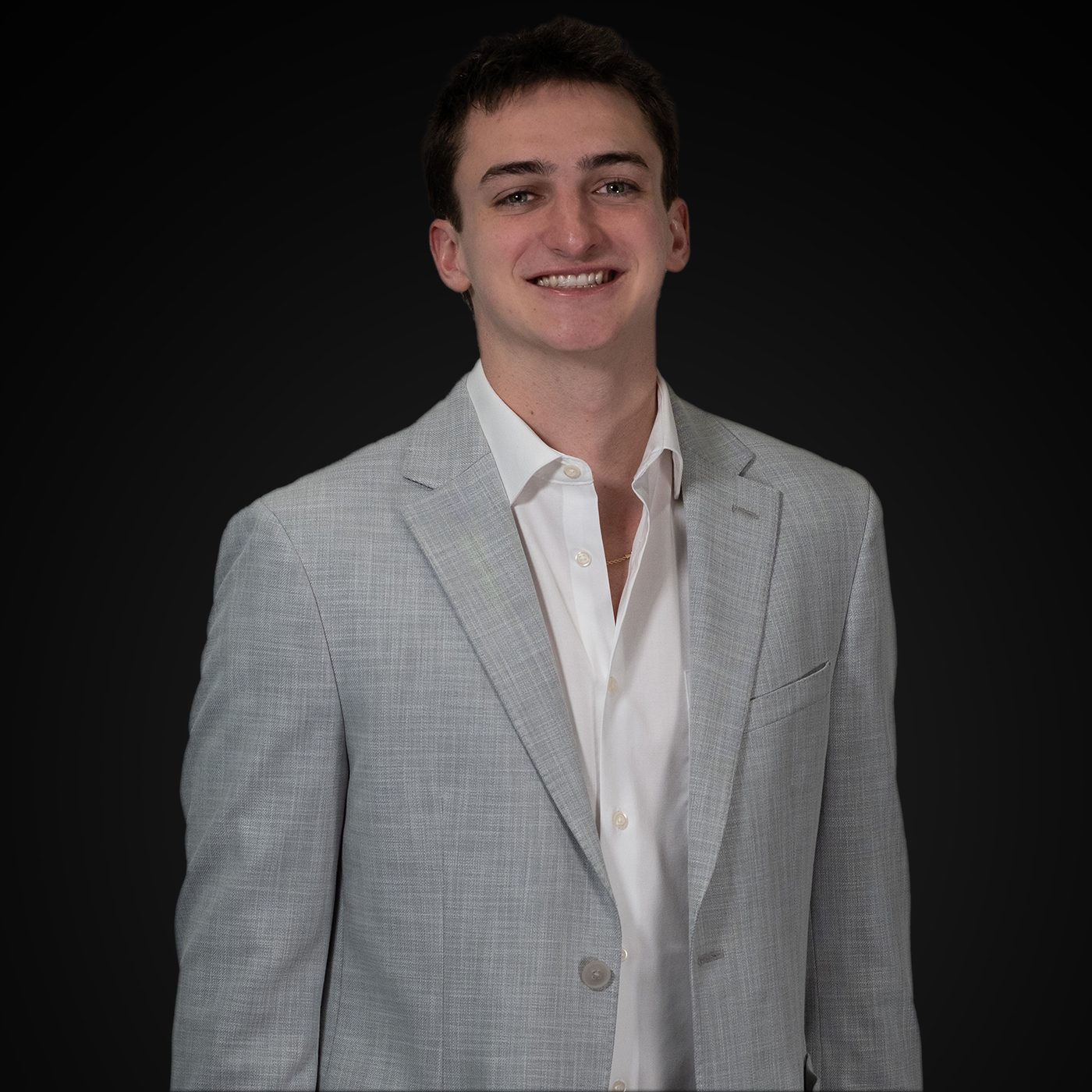 Young man in a gray blazer smiling against a black background.