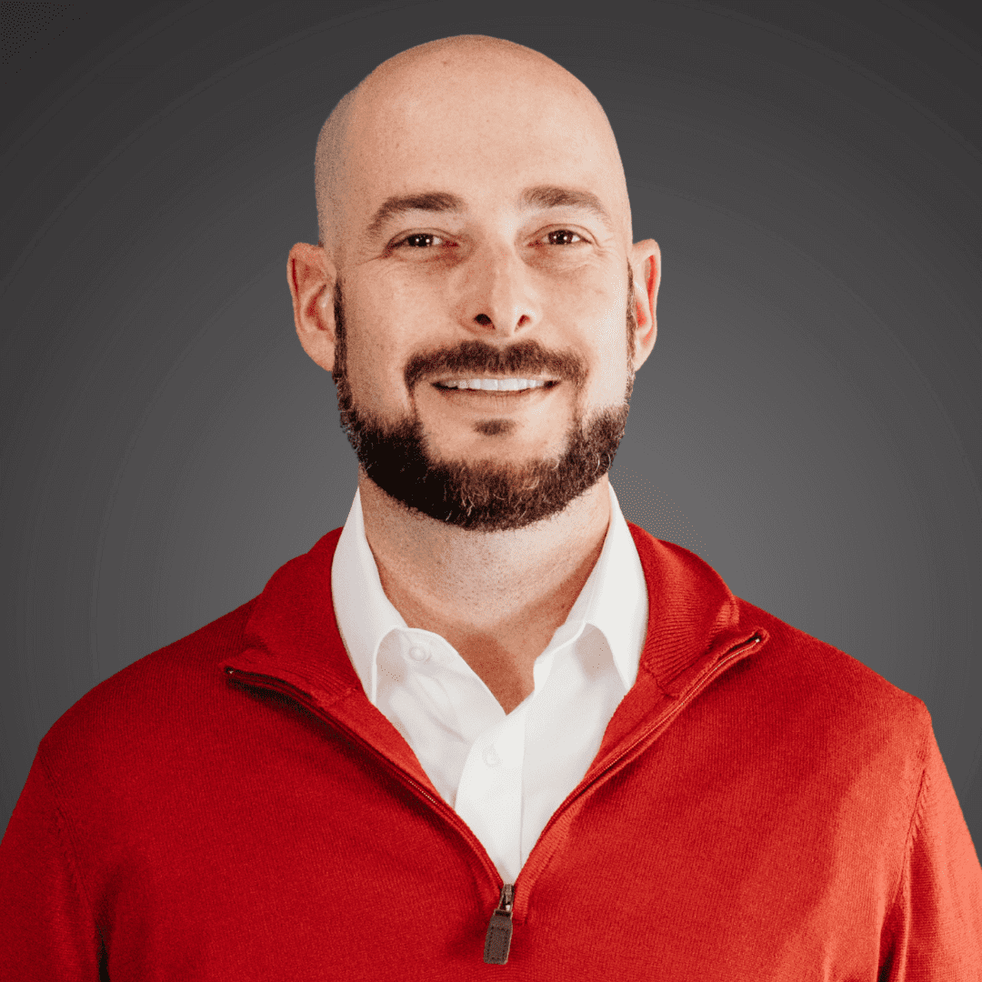 Smiling bald man with beard wearing a red jacket and white shirt.